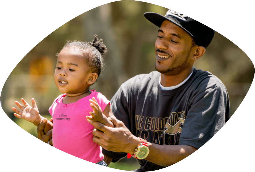 Smiling African Amercan father dressed in tee shirt and baseball hat is helping his smiling daughter stand